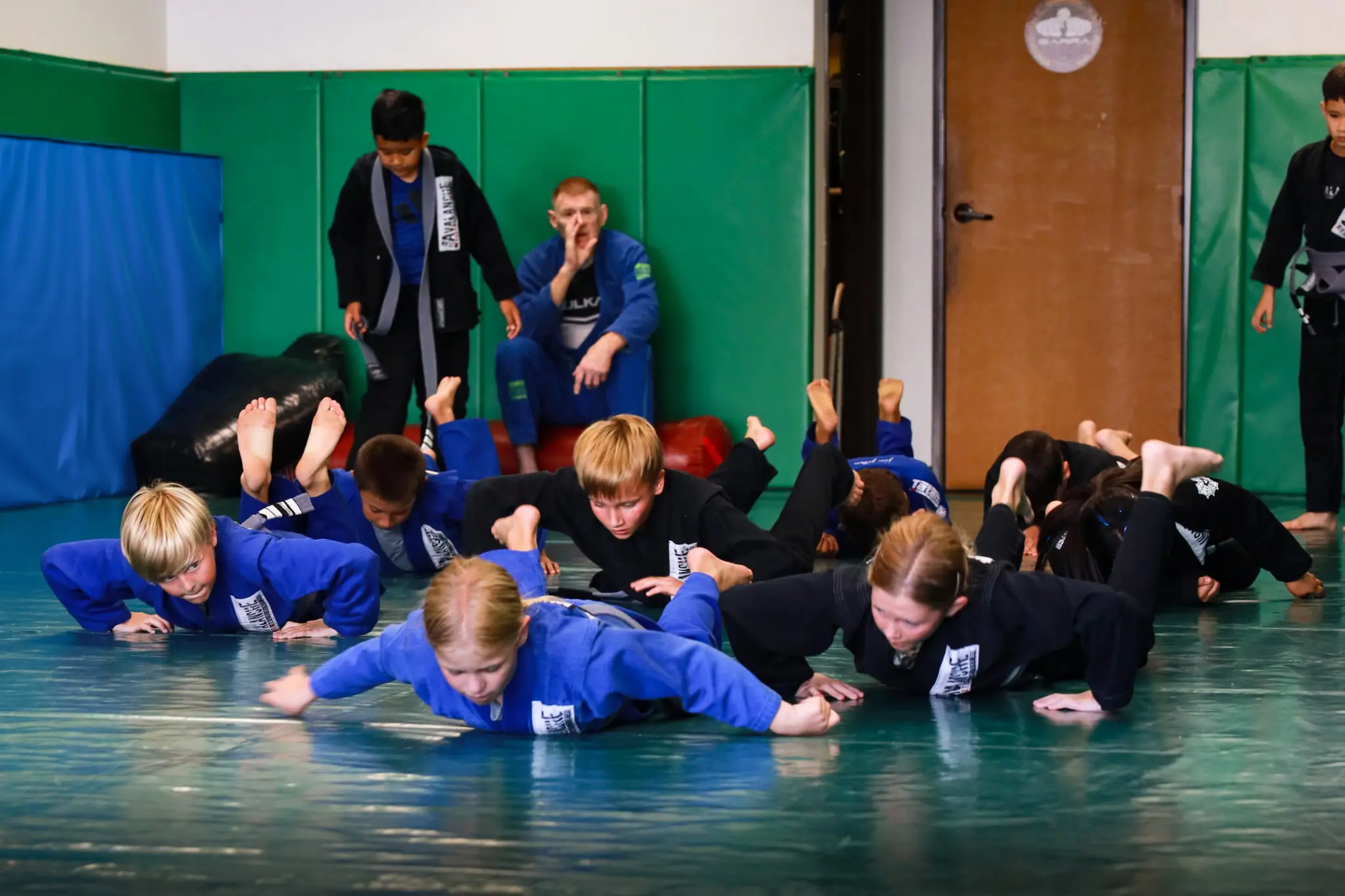 A full class of children holding a low position on the mat during a conditioning drill, with two coaches supervising