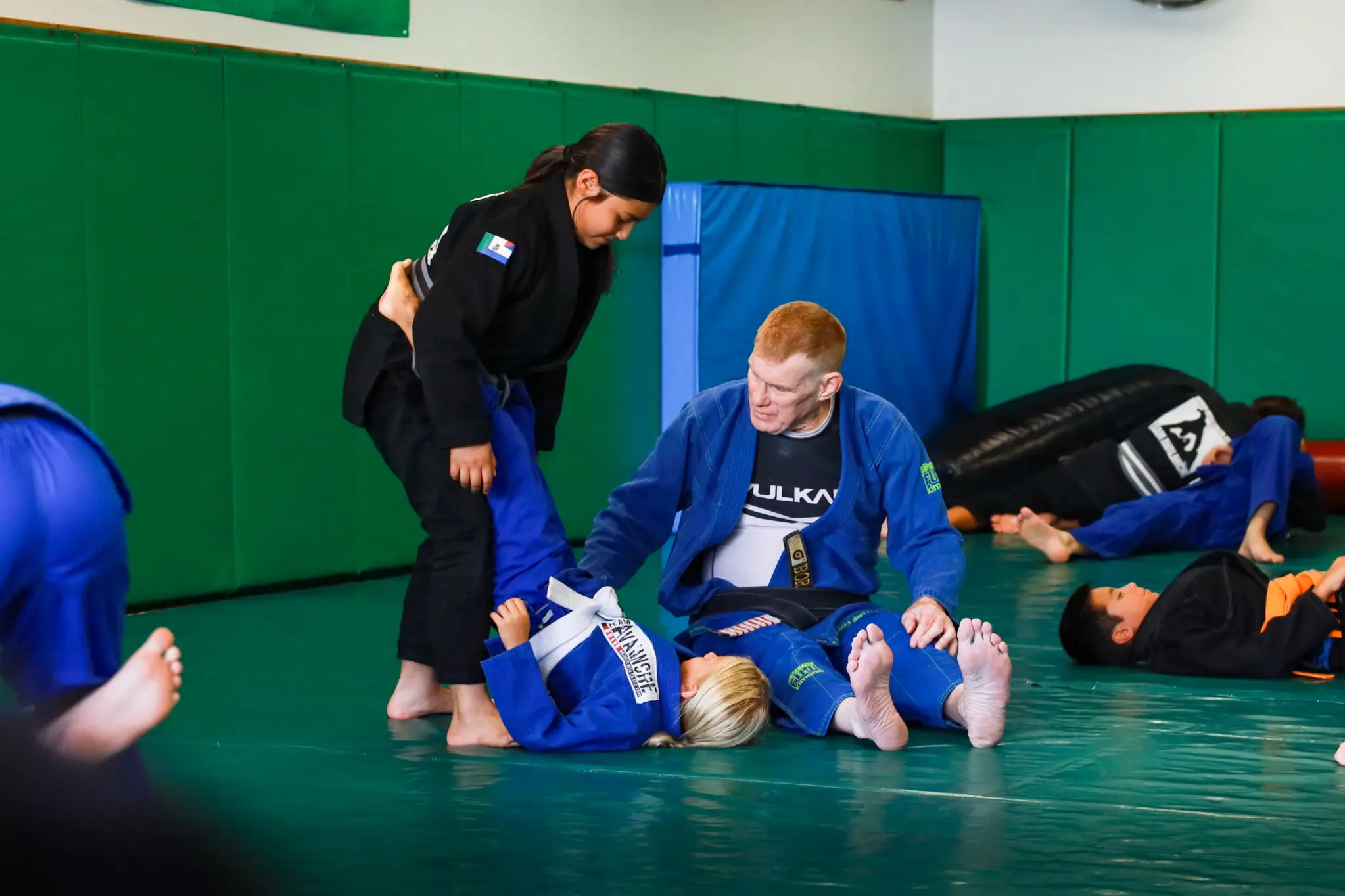 Professor James Boran kneeling beside two young students on the mat, correcting their position mid-drill