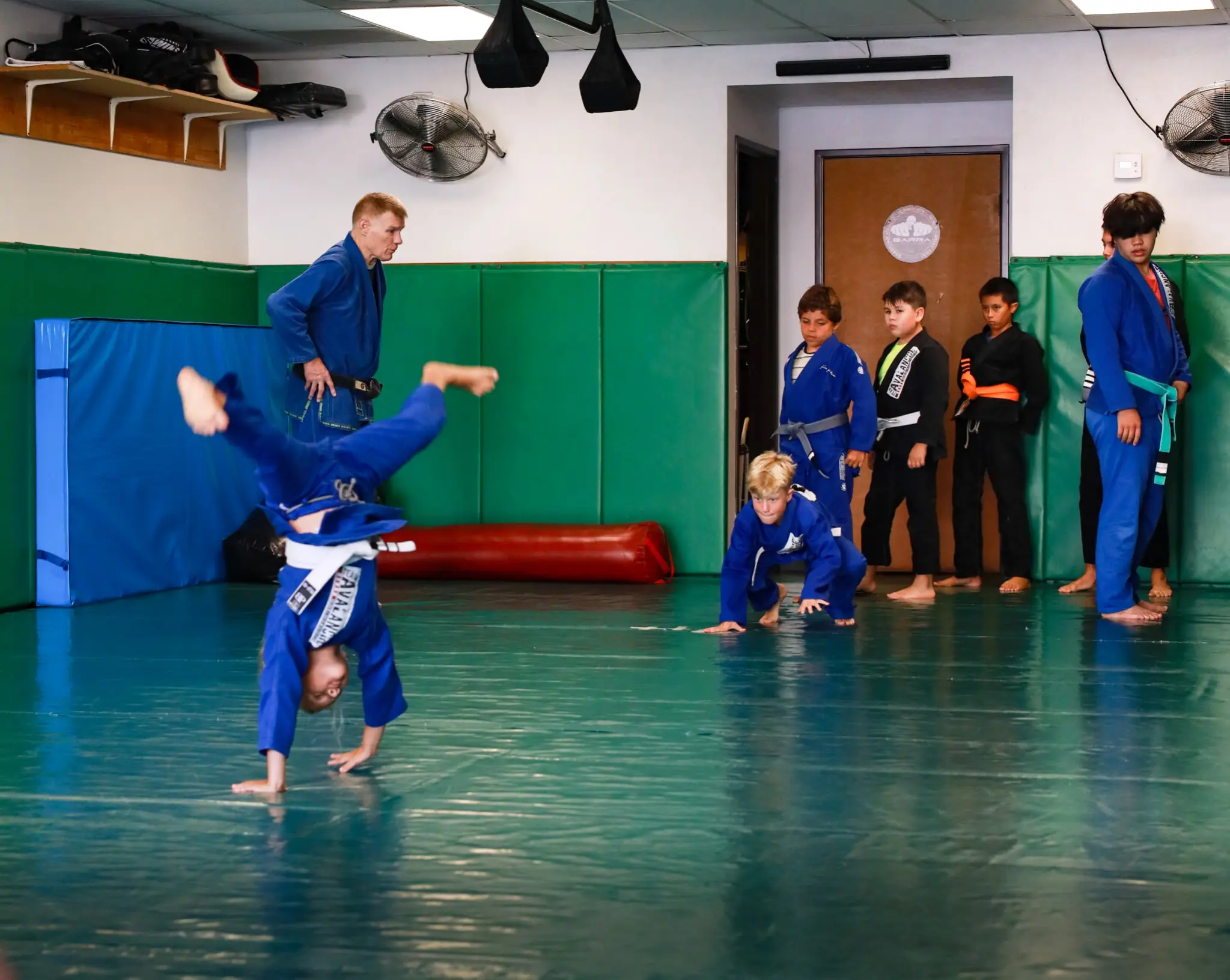 A child turning a cartwheel across the mat during a movement drill, watched by Professor James Boran while classmates wait their turn