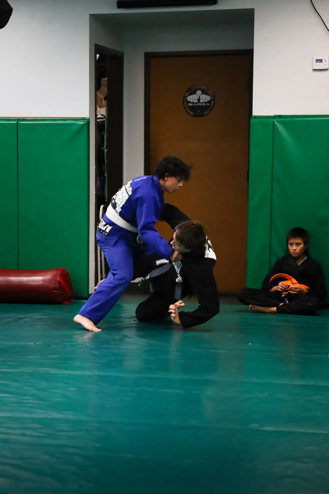 A student in a blue gi gripping the collar of a partner in a black gi who is working up from the mat, while a classmate waits by the door