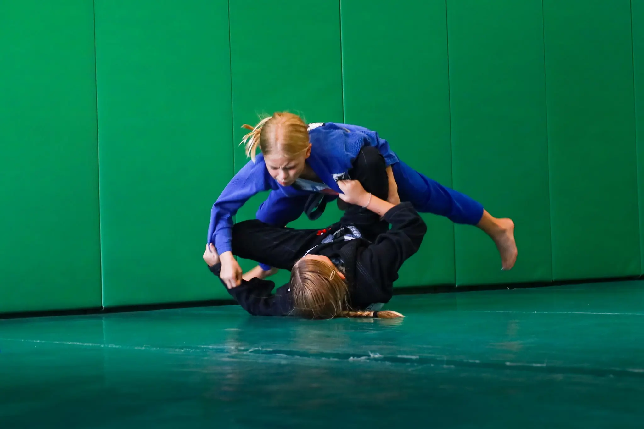 A young student in a blue gi diving past a partner’s guard during a live round