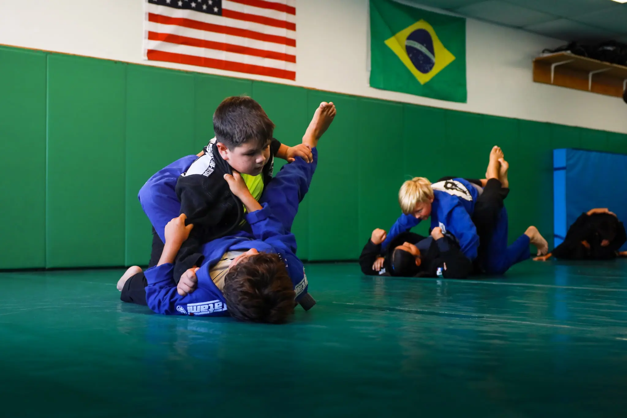 Two pairs of children rolling in the gi beneath the American and Brazilian flags at Boran Brazilian Jiu-Jitsu