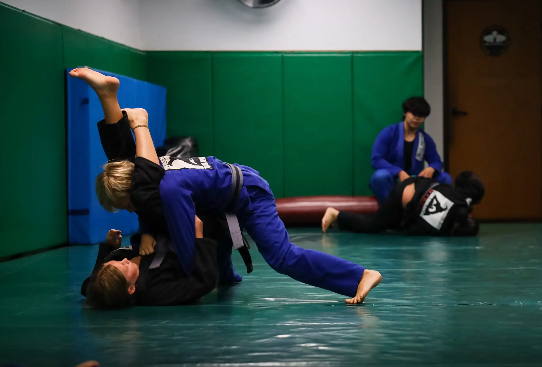 A young student in a blue gi driving past the raised legs of a partner in a black gi during a live round