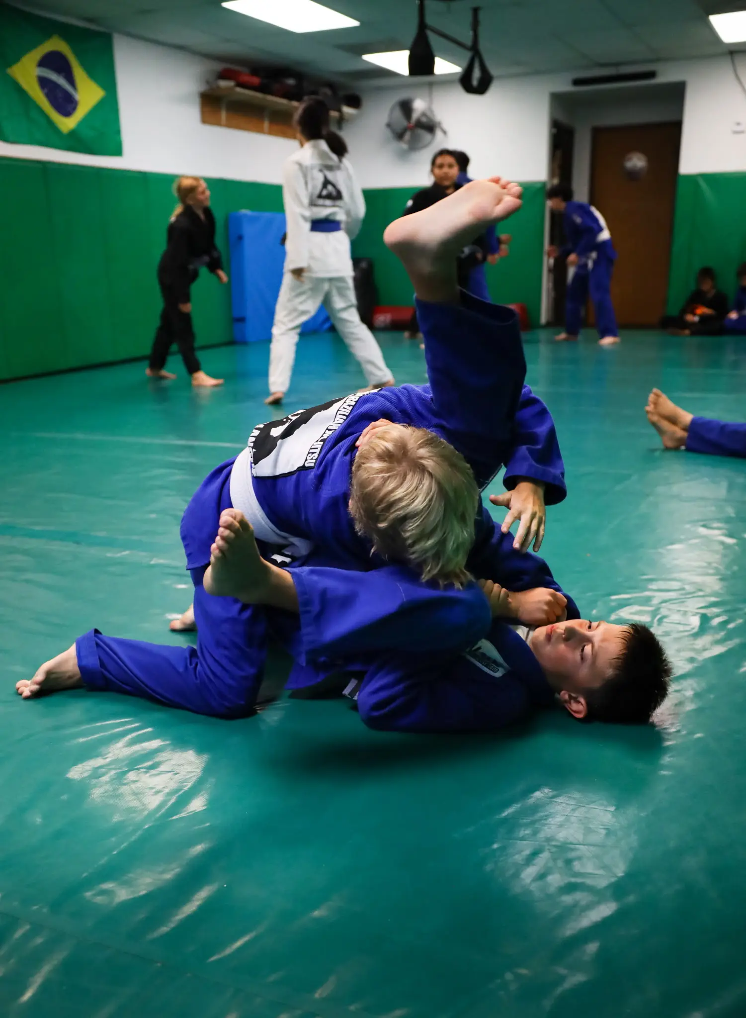 Two young students in blue gis in a scramble, one tumbling upside down over the other, beneath the Brazilian flag