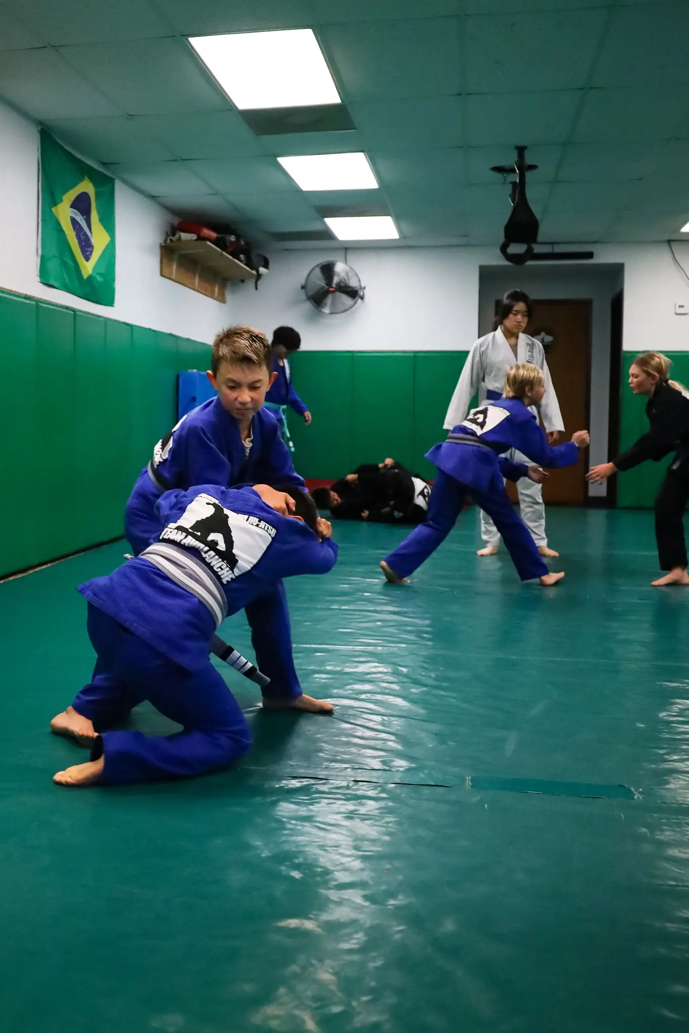 A young student on both knees driving into a standing partner for a takedown, with the class drilling behind them