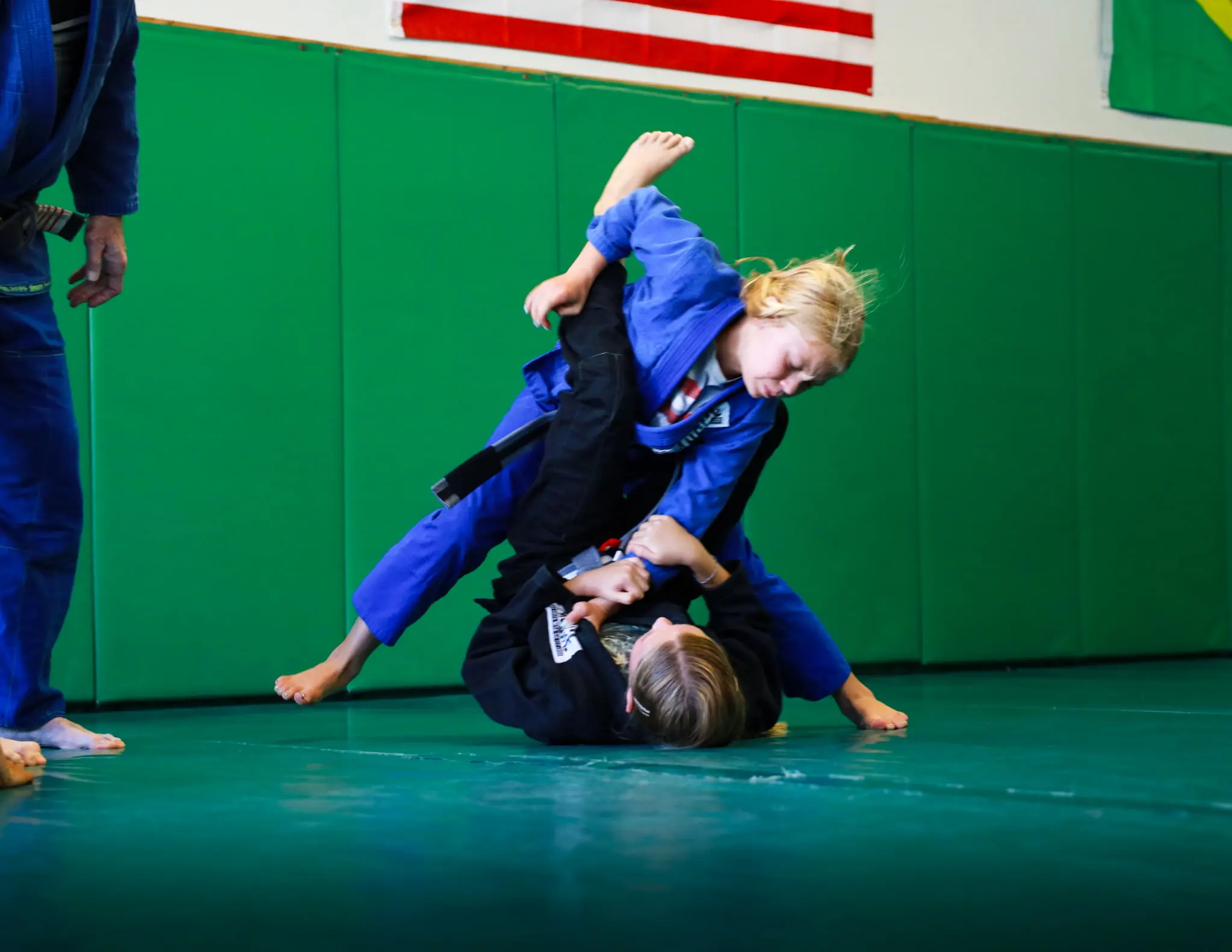 Two young students rolling in the gi, one holding top position, beneath the American and Brazilian flags