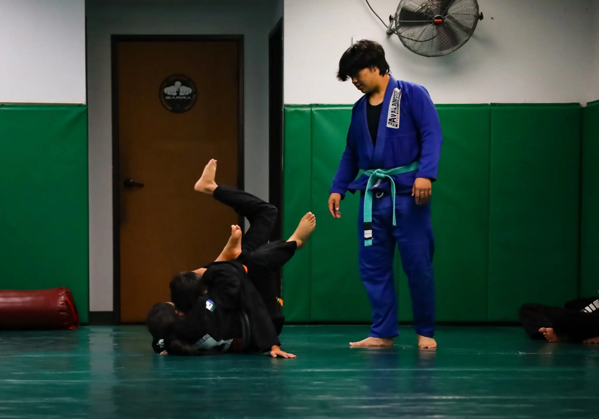 An older student in a blue gi and green belt looking on as two younger students in black gis roll beside him